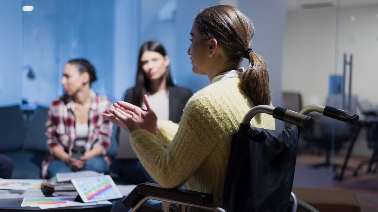 Young woman in wheelchair addressing two other young women seated on couch